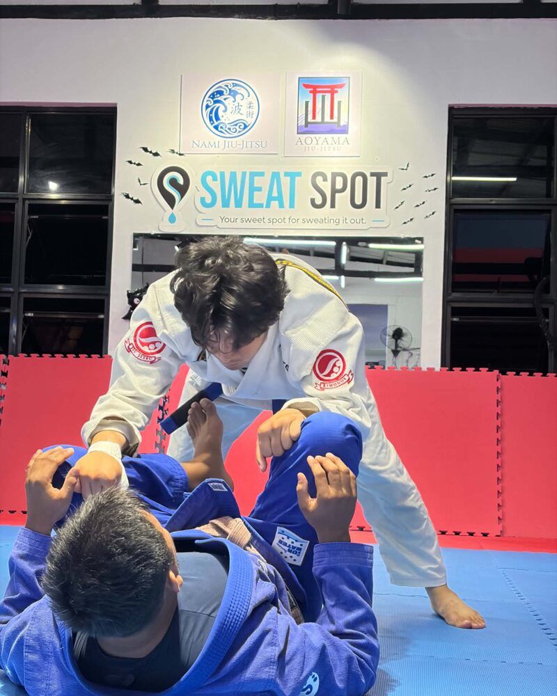Two athletes in gis rolling on blue mats, one standing and passing guard over the other lying on his back, beneath the Nami Jiu-Jitsu, AOYAMA Jiu-Jitsu and Sweat Spot signs